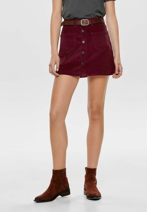 Femme portant une jupe en velours côtelé bourgogne à boutons sur le devant avec poches latérales, une ceinture marron, un haut noir et des bottes chelsea en suède marron.