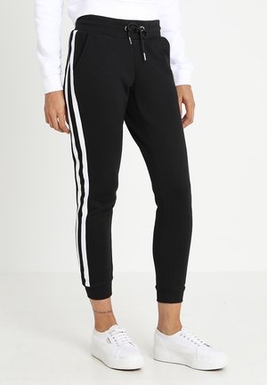 Person iført sorte joggerbukser med hvide sidelister og hvide sneakers, stående mod en ensfarvet lys baggrund.