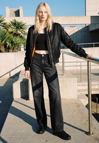 Šviesiaplaukė moteris stovi lauke ant betono laiptų, dėvi juodą trumpą palaidinę, juodą bomberio striukę, plačių kojų tamsius džinsus ir juodus sportinius batelius.