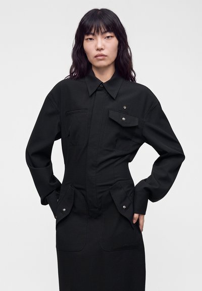 Femme aux longs cheveux foncés portant une robe noire à col avec des poches à rabat boutonnées, les mains posées sur les hanches devant un fond uni.