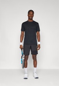 Černé sportovní tričko s krátkým rukávem, spárované s černými šortkami. Osoba drží tenisovou raketu, na nohou má bílé ponožky a tenisky.