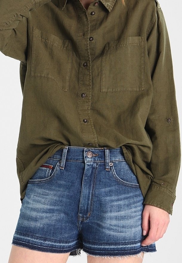 Camicia verde oliva a maniche lunghe con bottoni, dotata di due tasche sul petto, abbinata a pantaloni corti in denim blu scuro con orlo sfilacciato e una leggera scoloritura.