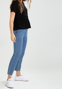 Top negro de manga corta con detalle de cintura con volantes, combinado con jeans ajustados azul claro y zapatillas blancas. Tejido suave con textura mínima.