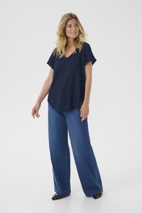 Tumši zila v-veida krekls ar īsām piedurknēm, ar teksturētu dizainu, apvienots ar platām džinsu biksēm un melnām norādītām kurpēm.