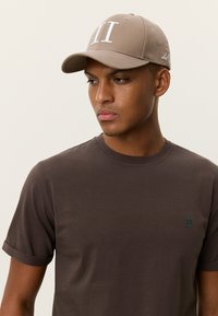 Μπεζ καπέλο μπέιζμπολ με λευκό κεντημένο λογότυπο "II" στο μπροστινό μέρος, φορεμένο από ένα άτομο με καφέ κοντομάνικο πουκάμισο με μικρό τιρκουάζ λογότυπο.