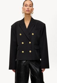Femme portant un blazer noir oversize à double boutonnage avec des boutons dorés et un pantalon large noir texturé, agrémentée de longues boucles d'oreilles en or.