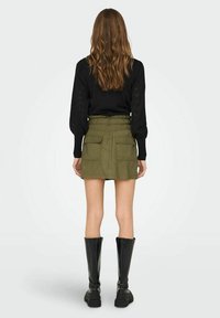 Jupe vert olive avec une taille ajustée, deux grandes poches et une coupe droite, associée à un haut noir à manches longues et des bottes noires jusqu'aux genoux.