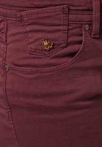 Pantalones burdeos con una textura suave, con un bolsillo delantero, botón dorado en forma de flor y detalles de costura a lo largo de las costuras.