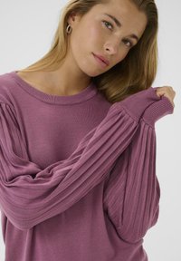 Mujer joven con cabello rubio, que lleva pendientes de aro y un suéter de manga larga de canalé morado, ajustando suavemente la manga sobre su mano.