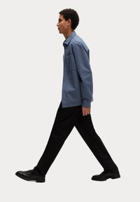 Uvolněná modrá košile na knoflíky s kapsou, spárovaná s černými kalhotami a černými botami, model jde v profilu na neutrálním pozadí.