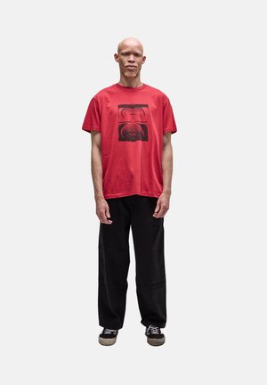Homme avec albinisme portant un T-shirt rouge à motif graphique, un pantalon noir et des baskets noires, debout devant un fond clair uni.