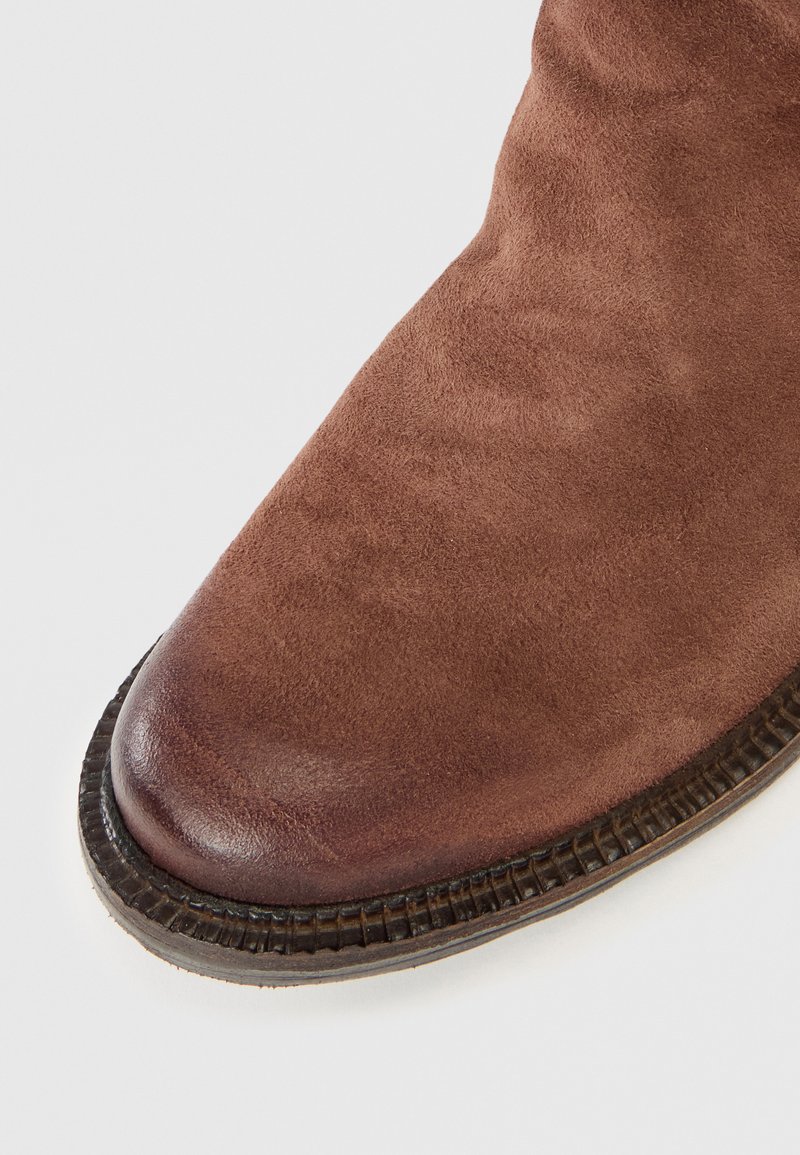 Bout en suède marron avec une semelle texturée marron foncé sur fond blanc, montrant des signes d'usure à l'avant.
