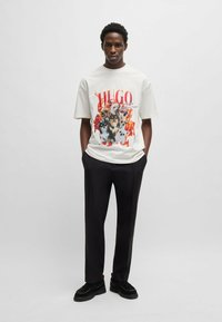 T-shirt bianco in cotone con una grafica colorata di cani e il nome del marchio "HUGO." Abbinato a pantaloni neri e scarpe nere spesse.