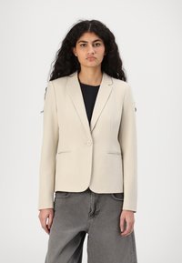 Beige bleiseri, joka on valmistettu sileästä kankaasta, yksinkertaisella nappisuljennalla, lovilliset läpät ja kaksi etutaskua, yhdistettyinä harmaisiin housuihin.