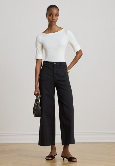 Femme debout en haut blanc ajusté, pantalon noir large taille haute, talons noirs ouverts, tenant un petit sac à main noir.