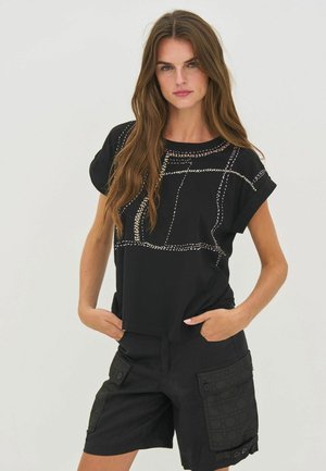 Mujer con cabello largo y castaño que lleva una camiseta negra de manga corta con patrones bordados y pantalones cortos cargo negros con grandes bolsillos.