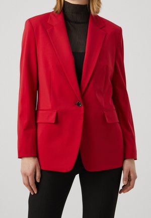 Žena nosící přiléhavý červený blazer s černým knoflíkem na průsvitném černém topu s vysokým límcem a černých kalhotách, stojící s uvolněnýma rukama po stranách.