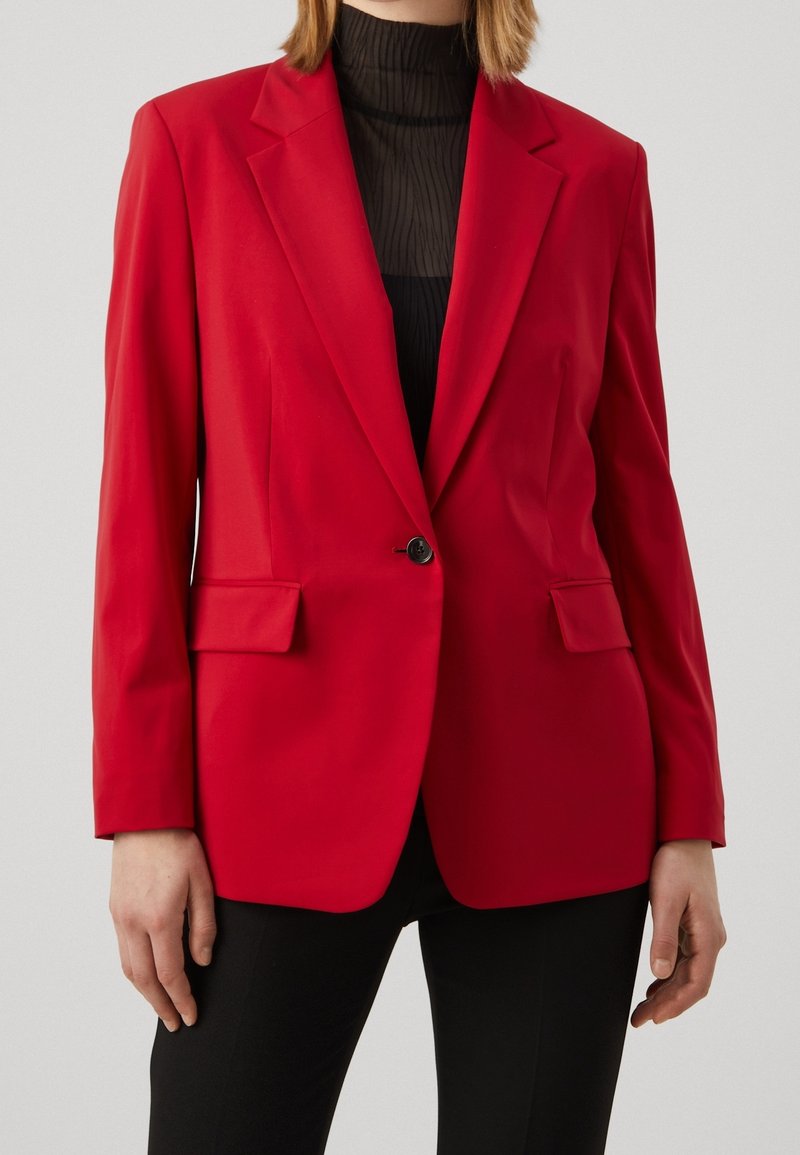 Femme portant un blazer rouge cintré avec un bouton noir sur un top noir transparent à col haut et un pantalon noir, debout, les mains relâchées sur les côtés.