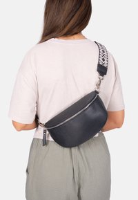 Schwarze Leder-Umhängetasche mit kurvenförmigem Design, ausgestattet mit einem Reißverschluss und einem gemusterten, verstellbaren Tragegurt. Texturierte Oberfläche für besseren Halt.