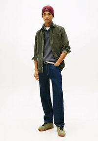 Olijfgroene corduroy shirt, grijze polo, donkerblauwe jeans, olijfgroene sneakers. Model draagt een kastanjebruine geribbelde beanie, met een borstzakdetail op het shirt.