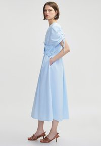 Robe midi bleu clair avec des manches courtes, taille smockée, détails floraux et poches latérales ; associée à des sandales à talons hauts à lanières marron.