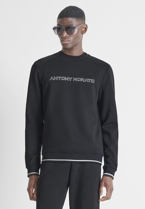 Uomo con felpa nera con la scritta "ANTONY MORATO", pantaloni neri coordinati e occhiali da sole scuri, in piedi davanti a uno sfondo chiaro.