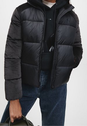 Doudoune noire avec des manches contrastées, fermeture éclair et détail de logo. Portée par-dessus un sweat à capuche sombre, assortie à un jean et tenant un sac.