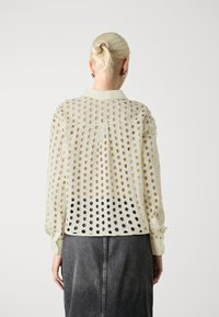 Lightweight, cream-colored blouse with large circular cutouts, long sleeves, and a buttoned collar, transitioning to black at the bottom.