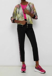 Kleurvolle zip-up jack met een bladmuster, gecombineerd met een felroze t-shirt en zwarte tapersl pants met zijsstripes; sportieve roze sneakers.