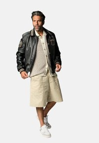 Veste varsity en cuir noir avec des accents crème, cardigan en tricot beige, shorts beiges et baskets blanches ; présente un patch sur la veste.