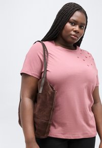 Donna con capelli lunghi intrecciati che indossa una maglietta rosa con piccoli dettagli a forma di cuore, che porta una borsa a tracolla in suede marrone.