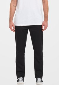Pantaloni di cotone neri con taglio dritto, caratterizzati da dettagli di cucitura discreti. Abbinati a una maglietta bianca e scarpe da ginnastica nere.