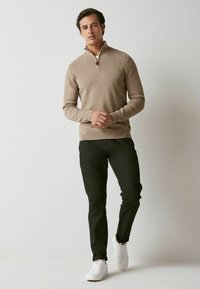 Beżowy sweter zapinany na zamek z teksturowanym wykończeniem, w połączeniu z ciemnozielonymi spodniami i białymi sneakersami, model stoi z złożonymi rękami.