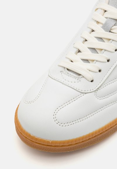 Hvite lær sneakers med glatt tekstur, beige gummisåle og flate hvite snøringer. Har minimal søm og en grå semsket detalj.