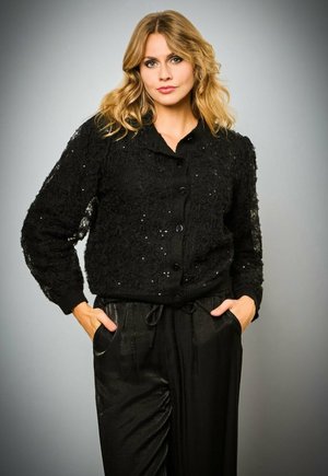 Zwarte kanten cardigan met bloemenpatroon, versierd met kleine pailletten, met een knoopsluiting en geribbelde manchetten. Gecombineerd met zwarte broeken.