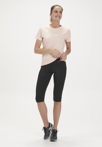 Ljusrosa tränings-T-shirt med vit text, kombinerad med svarta knälånga leggings. Modellen bär grå löparskor och står framför en neutral bakgrund.