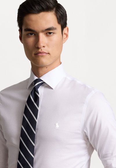 Chemise blanche à col avec un petit logo brodé sur la poitrine gauche, associée à une cravate rayée bleu marine avec des accents blancs.