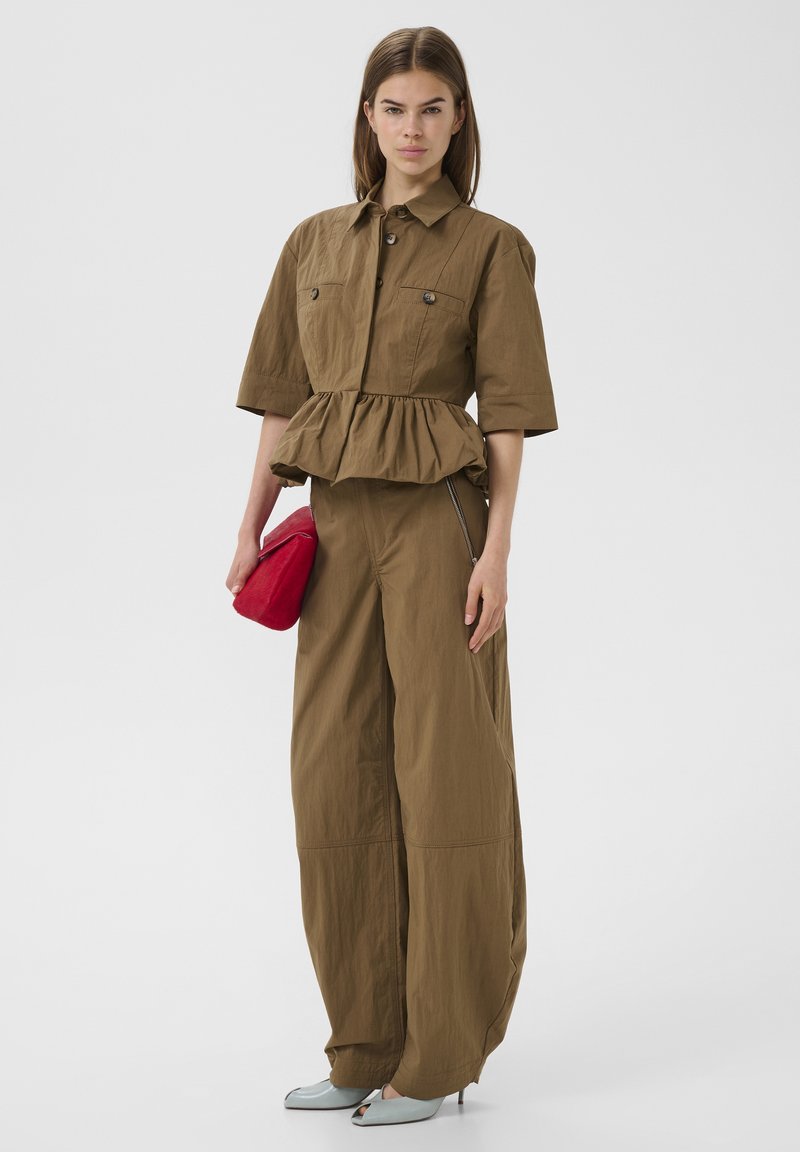 Vrouw met een bruin jasje met geplooide taille en wijde broekspijpen, een rode clutch in de hand, en lichtblauwe open-toe hakken, staande op een witte achtergrond.