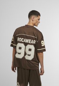 Barna, rövid ujjú mez, amelyen "ROCAWEAR" felirat és "99" szám található krémszínben. Kerek nyakkal és kontrasztos csíkokkal a ujjakon.