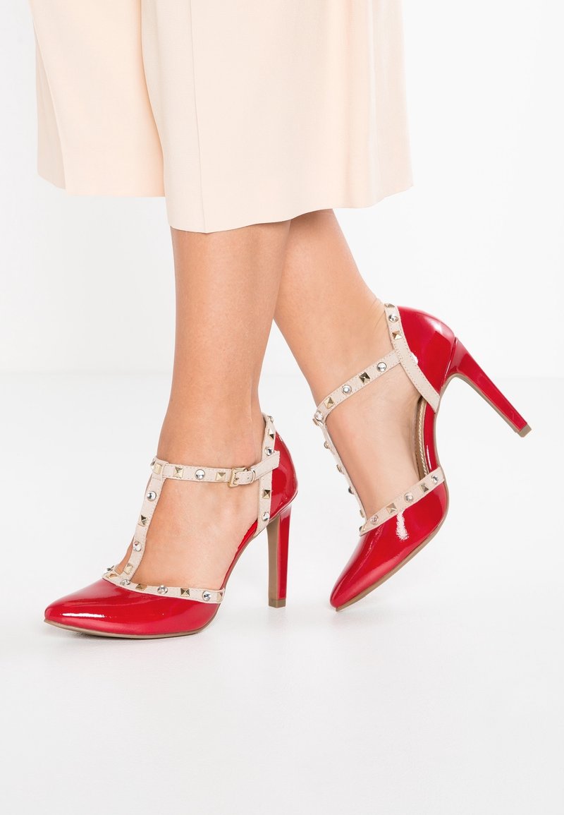 Chaussures à talons hauts en cuir verni rouge avec un bout pointu et des brides de cheville ornées de clous métalliques et d'un accent beige.