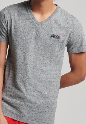 Mand iført en gråmeleret V-hals T-shirt med et lille "SuperDry"-logo på venstre bryst og røde bukser, mod en ensfarvet baggrund.