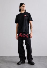 Black cotton T-shirt with logo patch; loose-fitting jeans in black with red cartoonish cat graphic on the back, yellow stitching, and textured footwear.