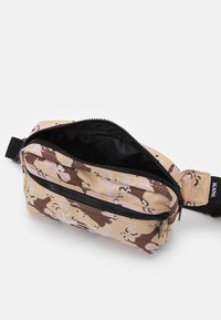 Hnědo-písková maskovací taška se zipovým uzávěrem, obsahující hlavní přihrádku a přední kapsu, vyrobená z texturované tkaniny.