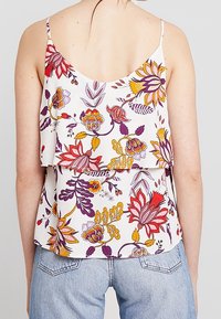 Femme portant un haut floral sans manches blanc avec des fleurs rouges, violettes et jaunes, associé à un jean bleu clair, vu de dos.