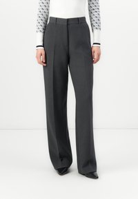 Donna che indossa pantaloni grigi scuri a vita alta e gamba larga abbinati a scarpe nere a punta e a un maglione bianco con maniche a fantasia.