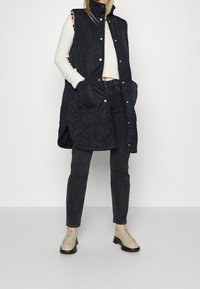 Gefütterte schwarze ärmellose Weste mit seitlichen Taschen, hohem Kragen und Druckknöpfen, getragen über einem weißen Pullover und dunklen Jeans.