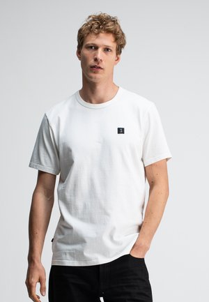 Jonge man met krullend blond haar, gekleed in een wit T-shirt met korte mouwen en een klein zwart logo op de borst, staande tegen een effen achtergrond.