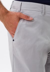 Pantaloni di cotone grigio chiaro con una texture liscia, dotati di una tasca frontale e un dettaglio con bottone sul lato. Design semplice e funzionale.