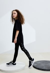 Črna ohlapna majica, črne legice z belimi vzorci in črni supergi z belimi podplati. Stoji na okroglih platformah, svetla ozadje.