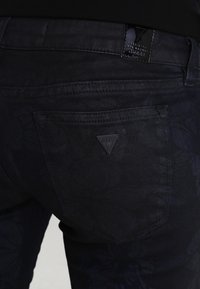 Jeans de mezclilla oscura con un sutil patrón floral, con un bolsillo trasero visible y un parche de cuero negro estampado con el logo de la marca.